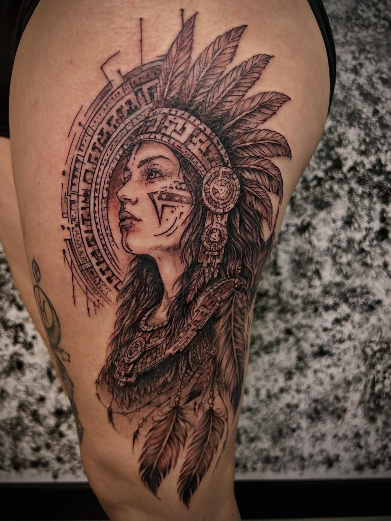 Tatouage réaliste noir et gris représentant le portrait d'une femme autochtone avec coiffe de plumes et motifs géométriques, réalisé sur la cuisse chez KavaDerma à Drummondville.