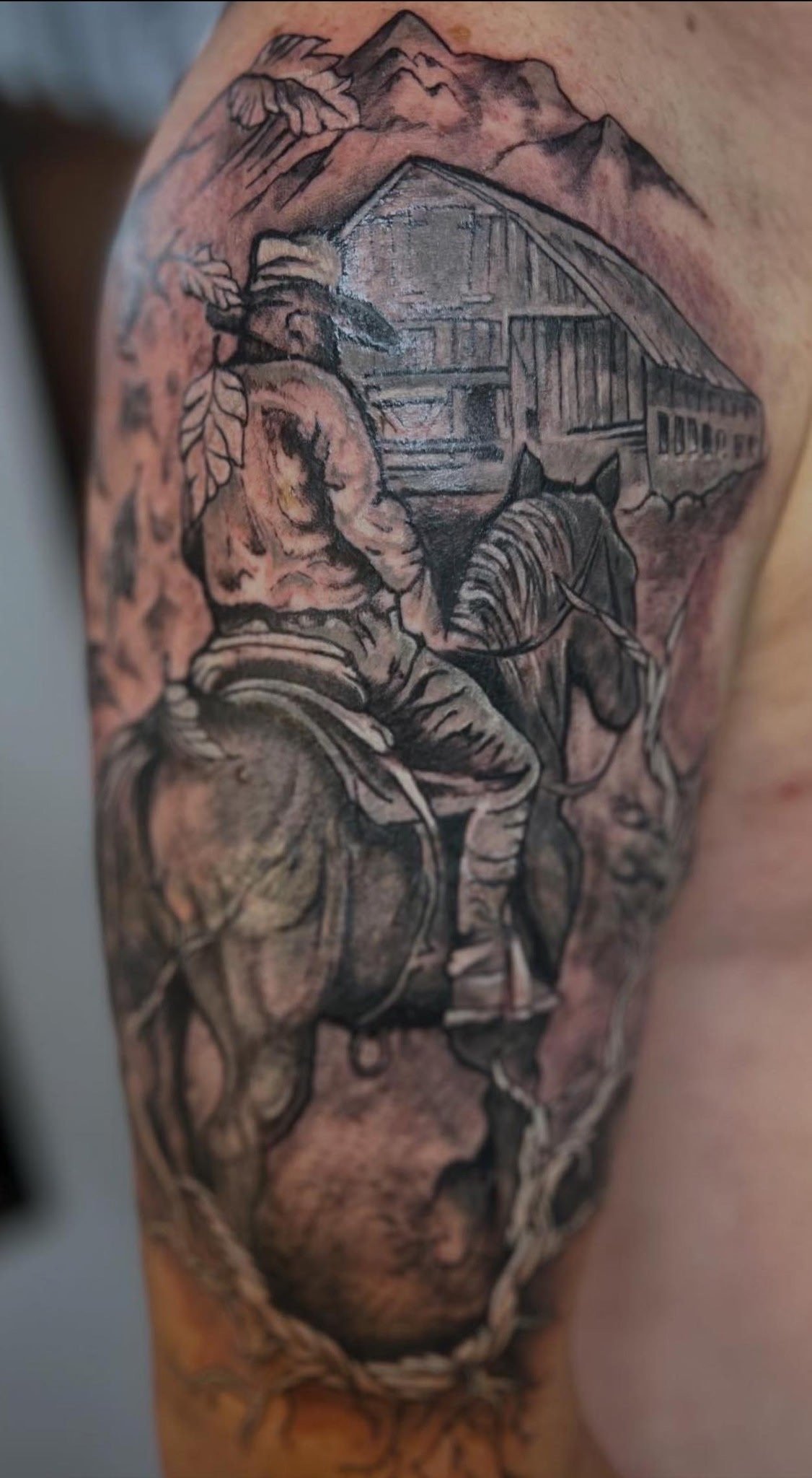 Tatouage réaliste noir et gris représentant un cowboy à cheval devant une grange avec des montagnes en arrière-plan, réalisé sur le bras chez KavaDerma à Drummondville.