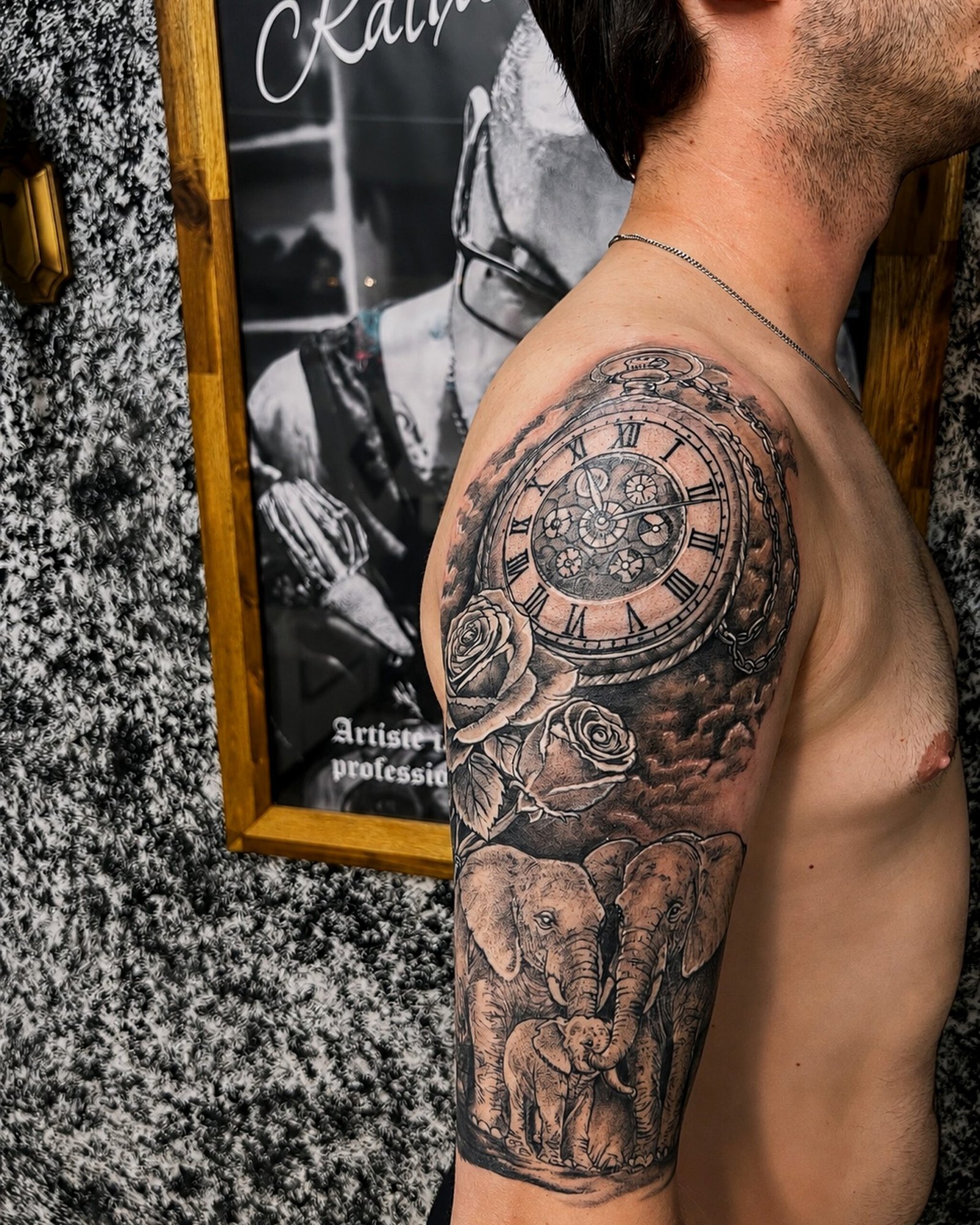 Tatouage réaliste noir et gris représentant une montre à gousset, des roses et une famille d'éléphants sur l'épaule et le bras, réalisé chez KavaDerma à Drummondville.